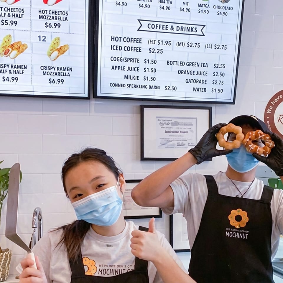 Masked bakery staff member holds a pretzel-like pastry while a colleague gives a thumbs-up behind the counter.