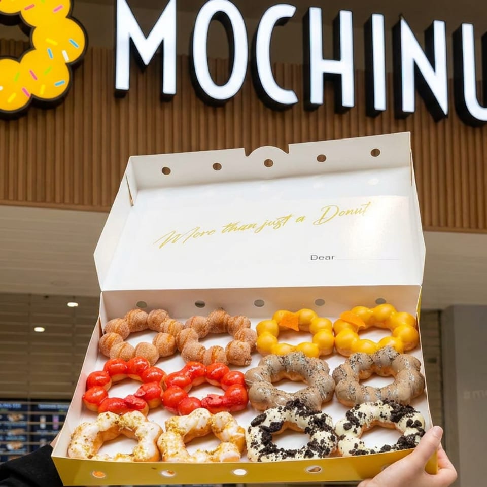 Open box of assorted colorful ring donuts held in front of a Mochinut storefront sign.