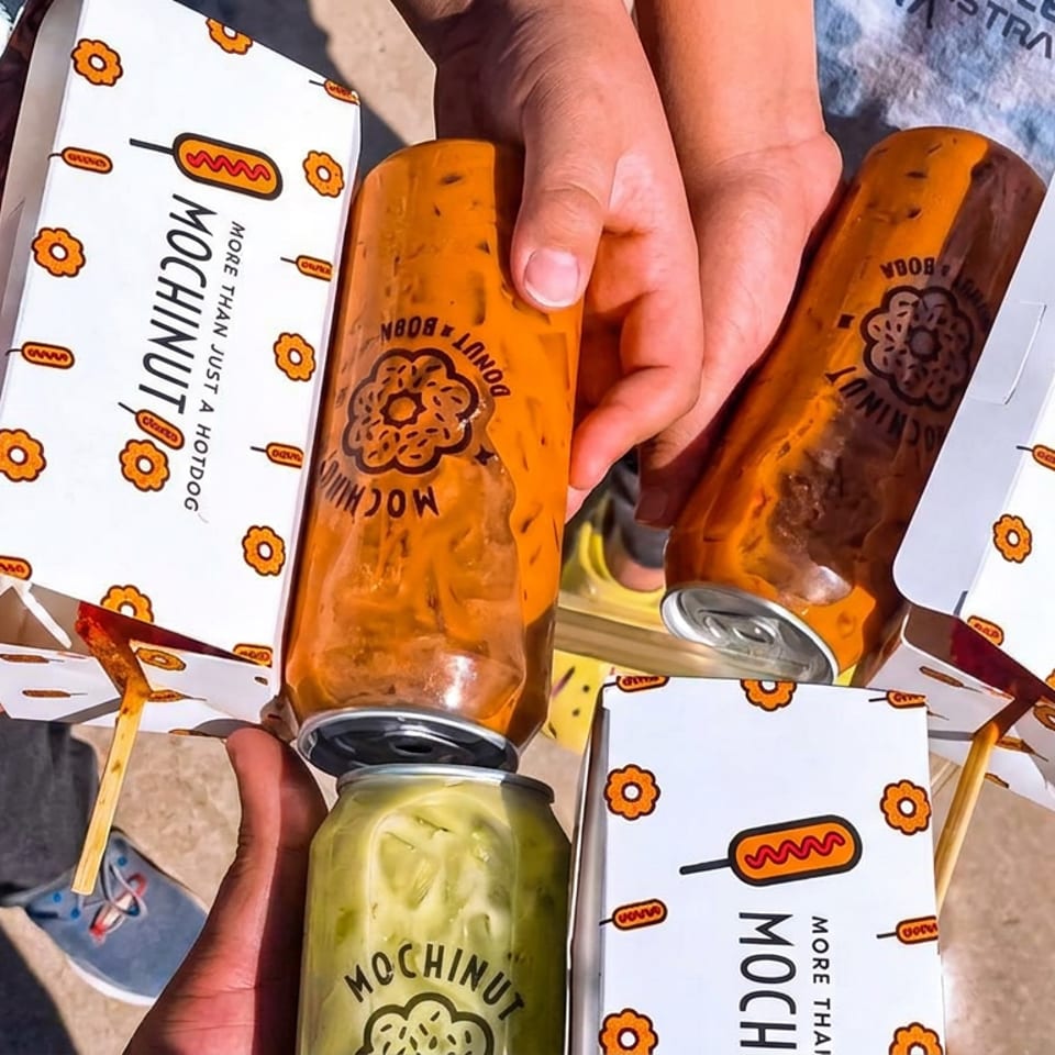 People hold colorful Mochinut hot dog boxes and drinks outdoors in bright sunlight.