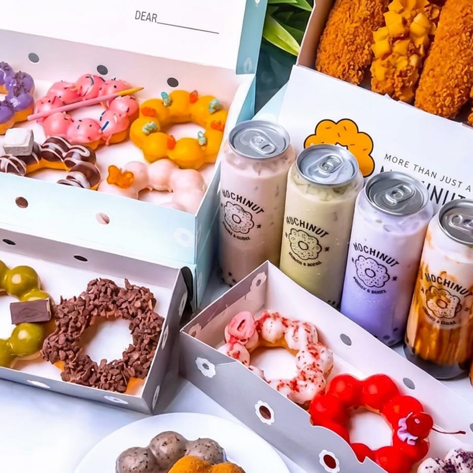 Open boxes display colorful decorated doughnuts, fried chicken strips, and assorted Mochinut drinks arranged outdoors against greenery.