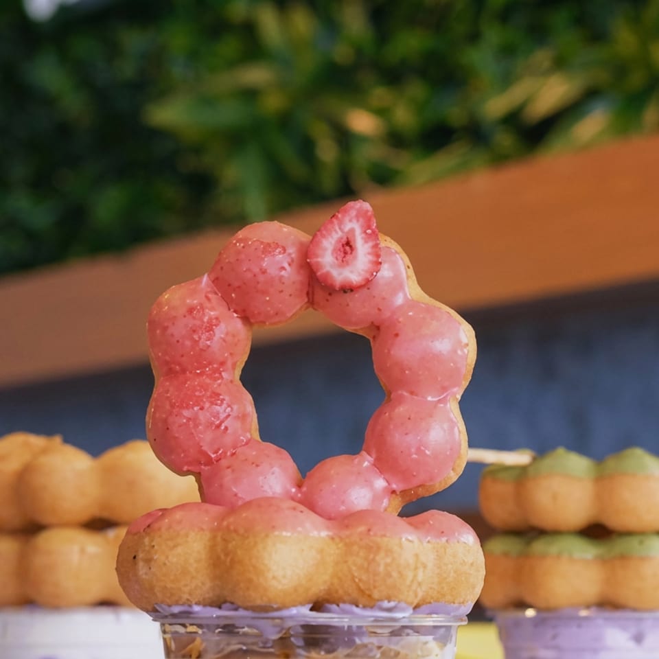 Pink strawberry-glazed ring donut topped with a strawberry sits on a clear cup, flanked by assorted donuts in the background.