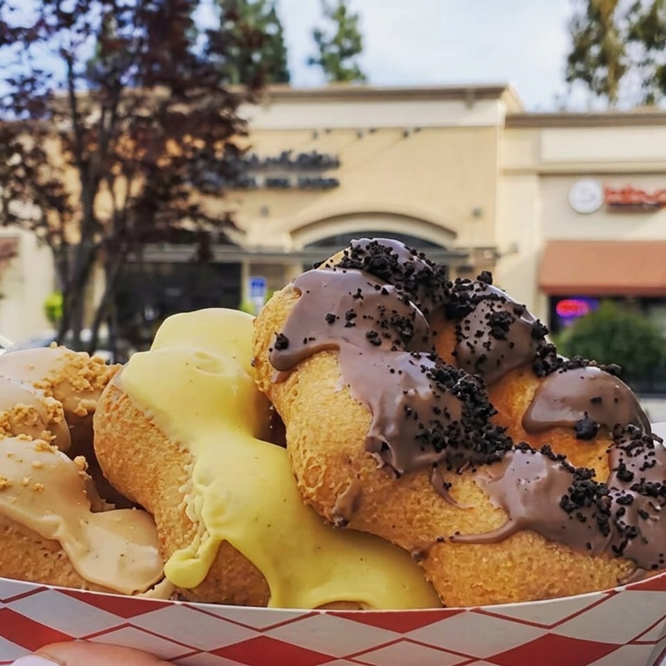 Basket of golden fried pastries topped with vanilla and chocolate icing and cookie crumbs outside a shopping-center storefront.