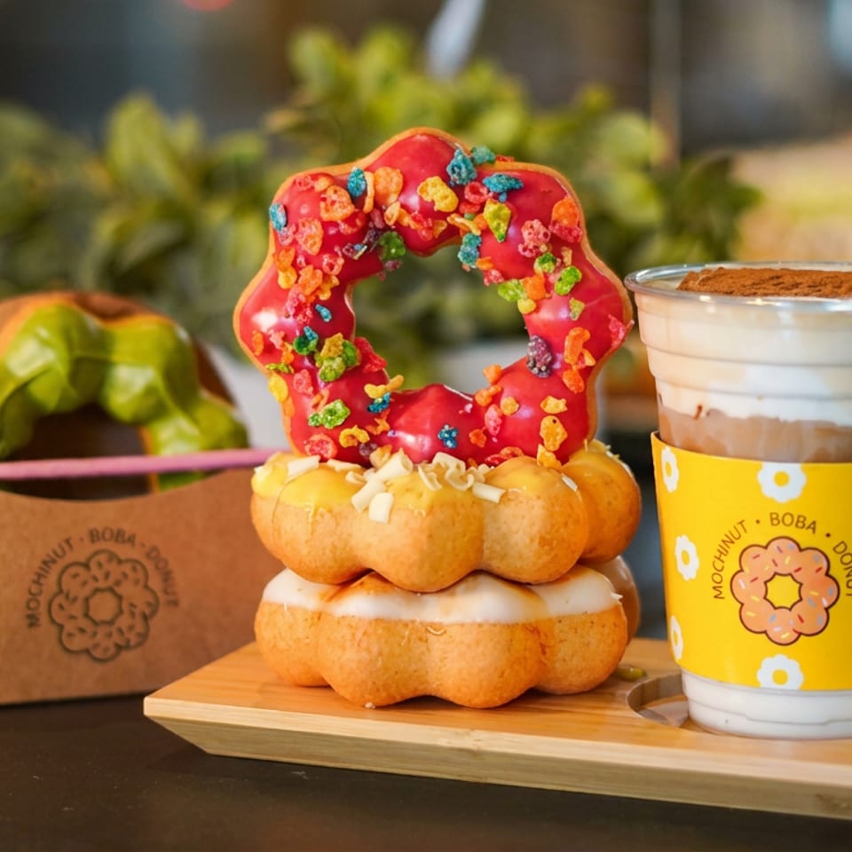 Colorful Mochinut doughnuts, including a red sprinkle-covered ring, sit beside a cup of boba tea and takeout box.
