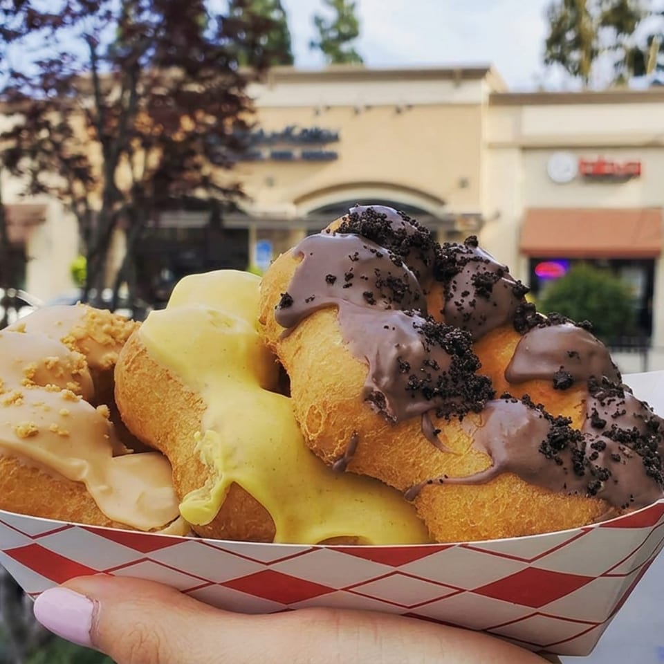 Basket of assorted glazed doughnuts topped with cookie crumbs, held outdoors near a shopping center.