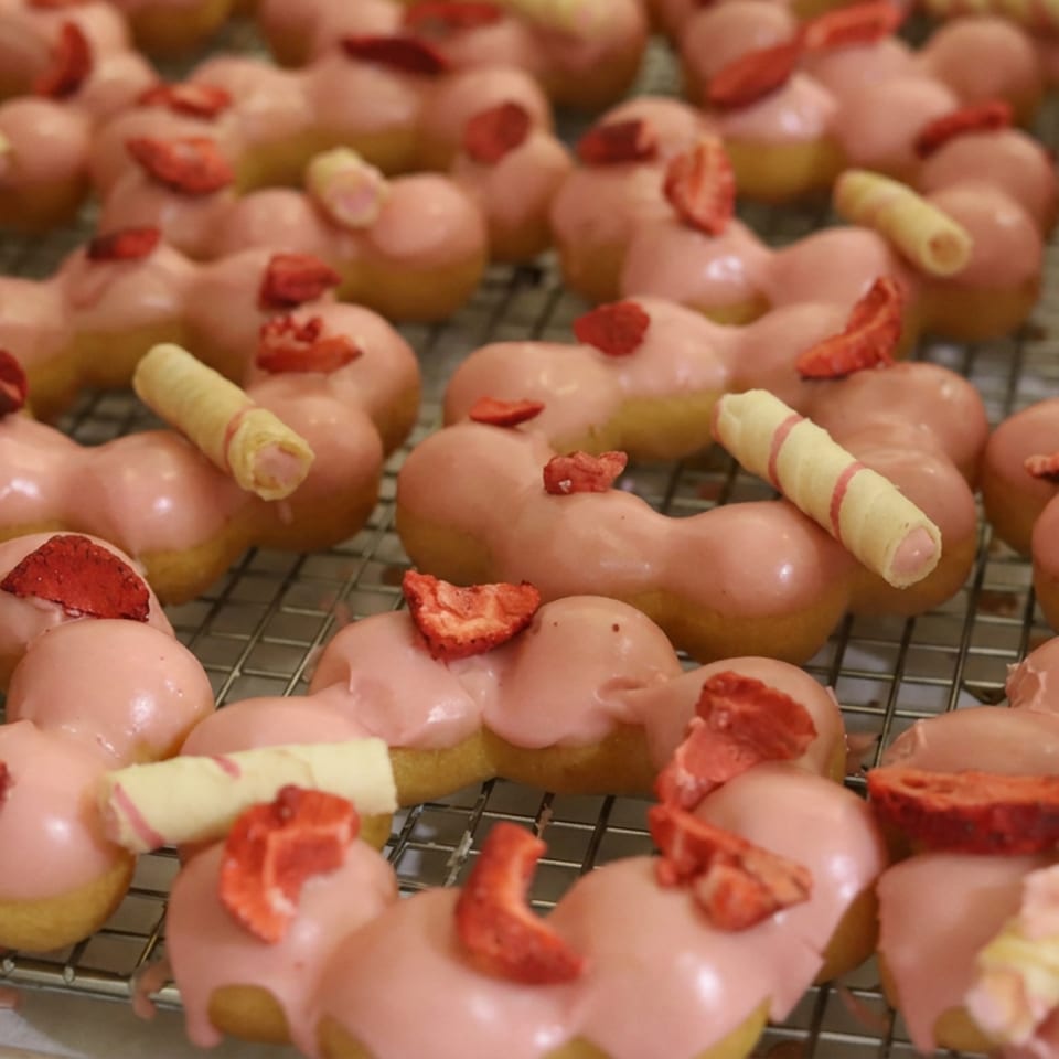 Rows of heart-shaped pastries with pink glaze, dried strawberry pieces, and wafer rolls cooling on a wire rack.