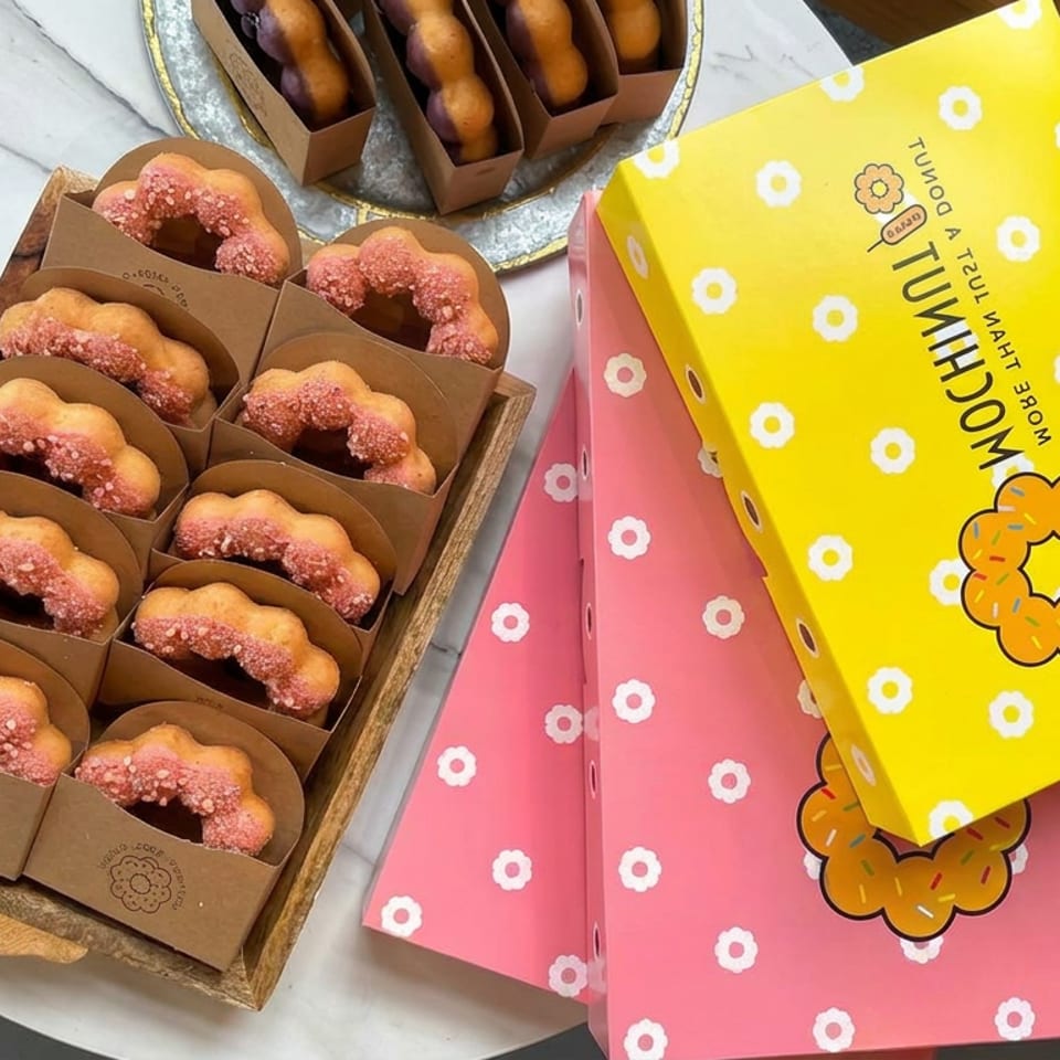 Assorted flower-shaped donuts with pink frosting in trays beside colorful floral boxes and a decorative flower arrangement.