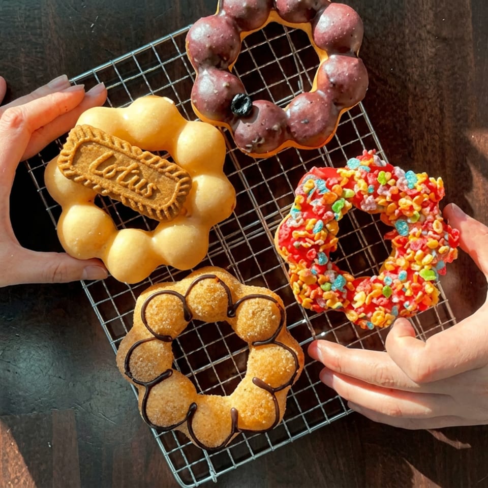 Four colorful ring-shaped donuts, including a berry-glazed, cereal-covered, chocolate-drizzled, and branded yellow donut, on a cooling rack.