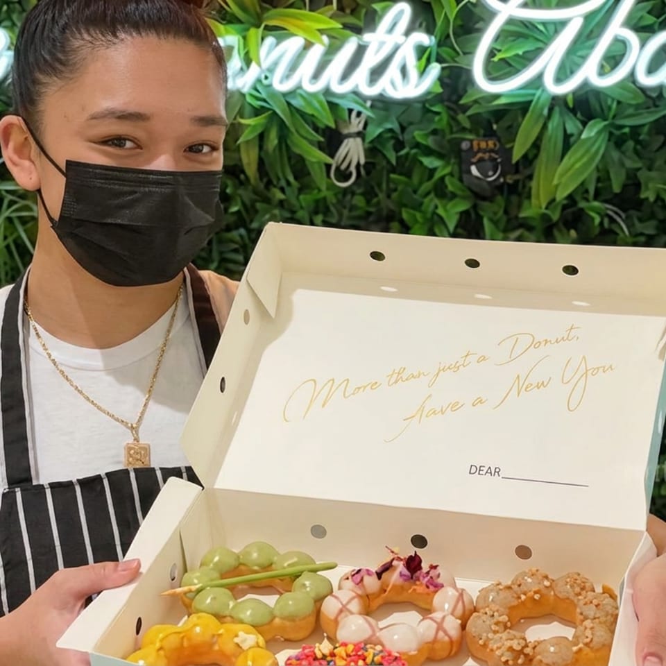 Masked bakery employee holds an open box of colorful decorated donuts against a leafy wall with a glowing sign.
