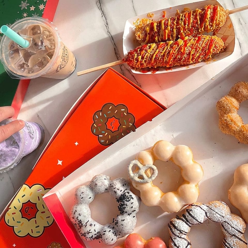 Mochi donuts, Korean corn dogs drizzled with sauces, and iced drinks arranged on a marble table.