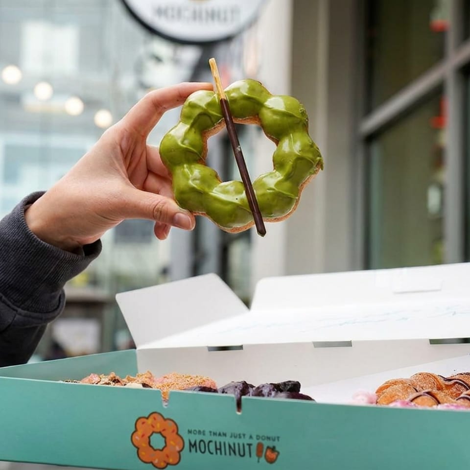 Hand holds a green matcha-glazed mochi donut above an open box of assorted Mochinut donuts on a city street.